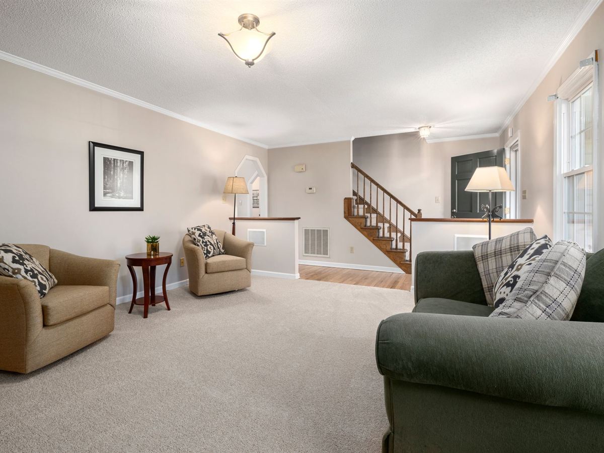 Living room with wall-to-wall beige carpet, tan armchairs and a staircase, lamps lit