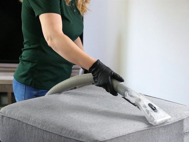 Gloved hands drawing an upholstery extraction wand across a grey fabric cushion