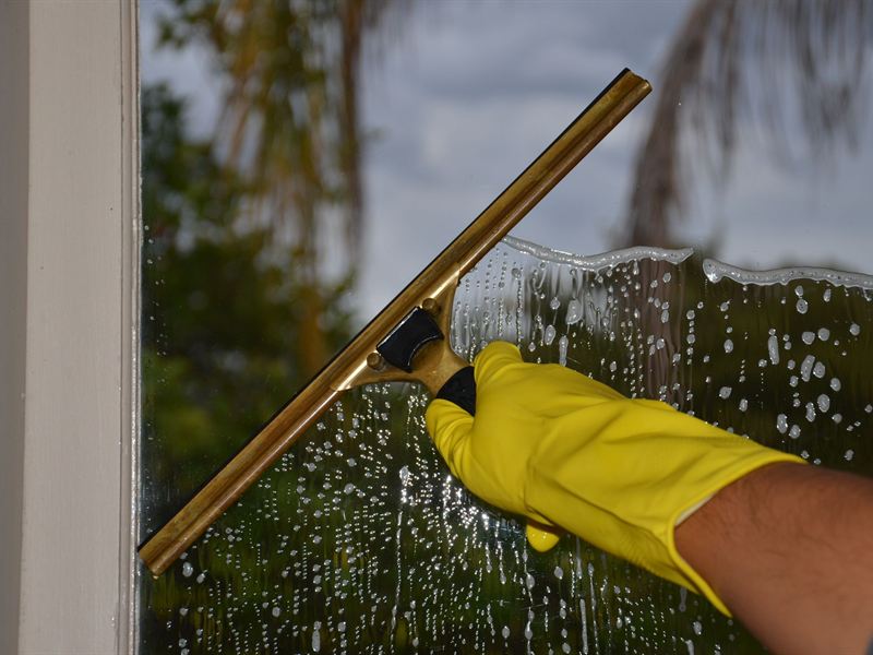 A gloved hand pulling a squeegee down a soapy window pane in a white timber frame